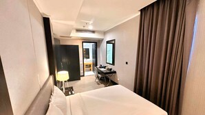 A hotel room with a large bed, a desk with a chair, a desk lamp, a mirror, and a door leading to a bathroom. - Le Charmé Suites (Olongapo)