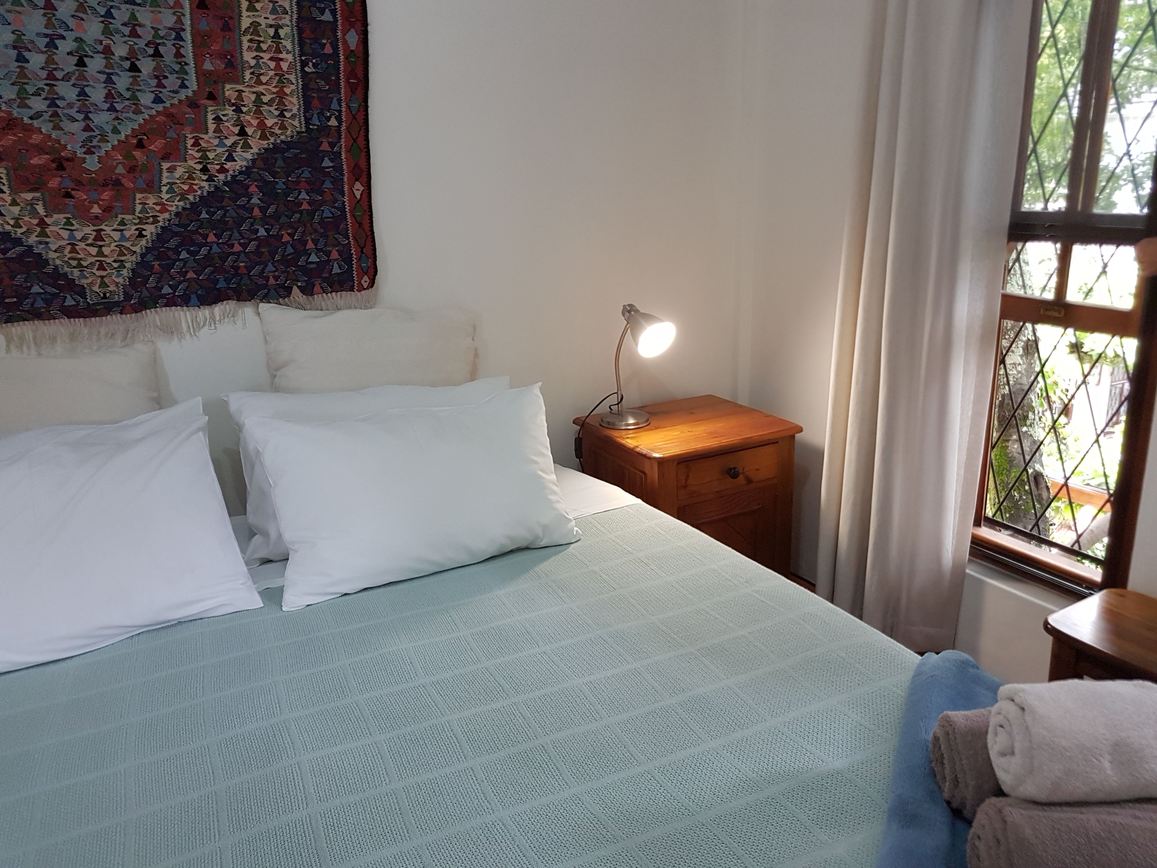 A neatly made bed with white pillows and a light blue bedspread, a wooden nightstand with a lamp, and a window with a view of greenery.