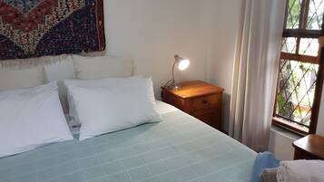 A neatly made bed with white pillows and a light blue bedspread, a wooden nightstand with a lamp, and a window with a view of greenery.