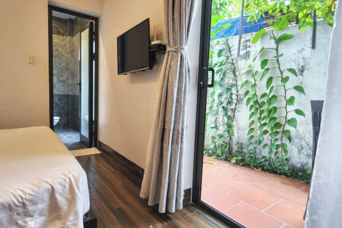 A hotel room with a bed, a TV mounted on the wall, a curtain, and a view of a garden with plants.