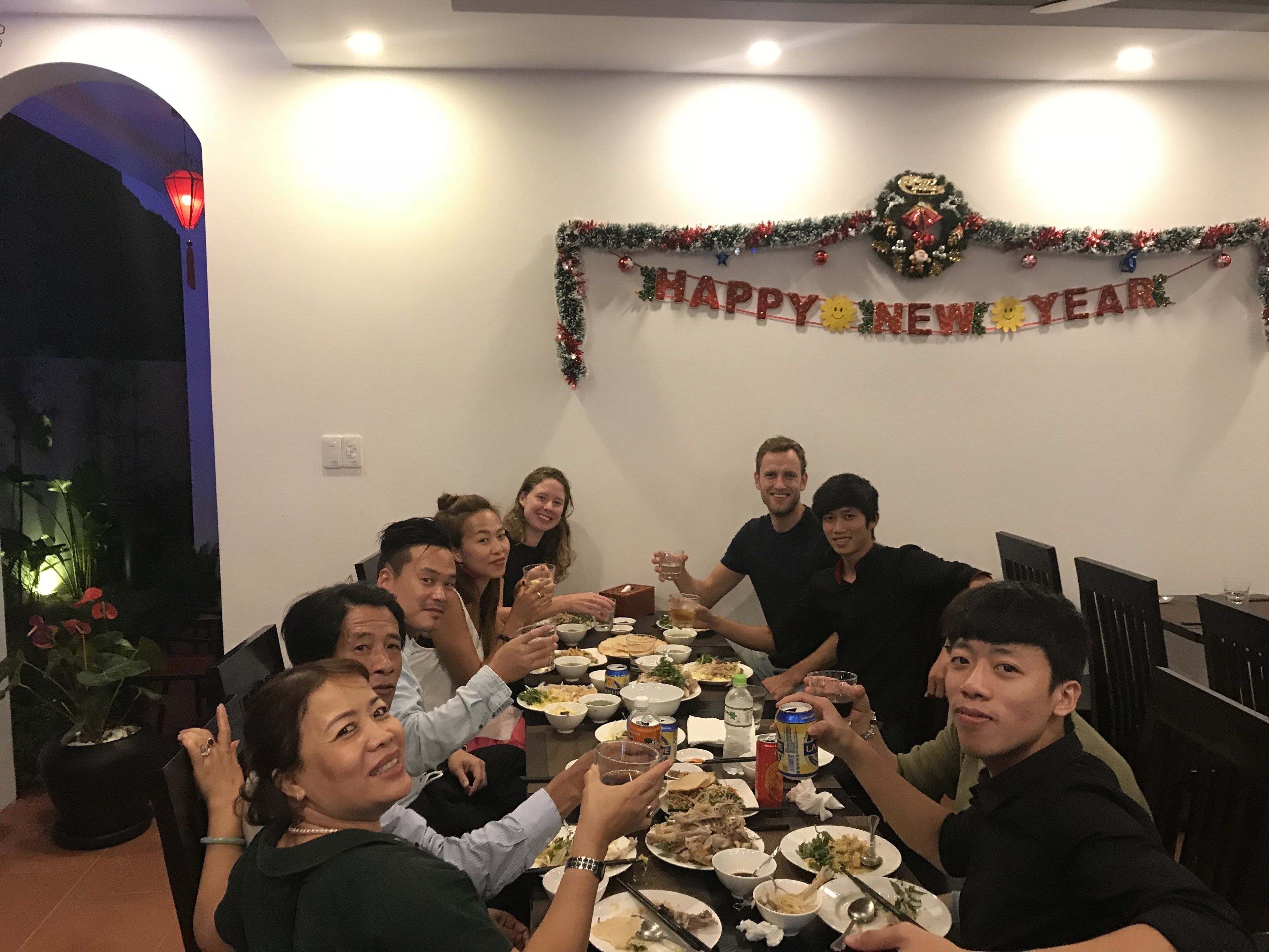 Un grupo de personas sentadas alrededor de una mesa, con un cartel que dice 'Happy New Year', indicando una celebración.