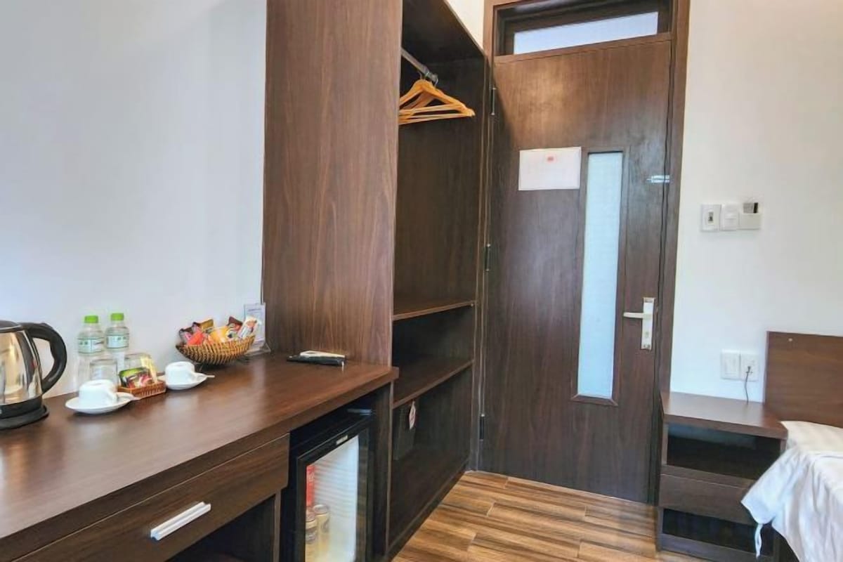 A compact hotel room with a wooden desk, a small refrigerator, a wardrobe, and a bed.