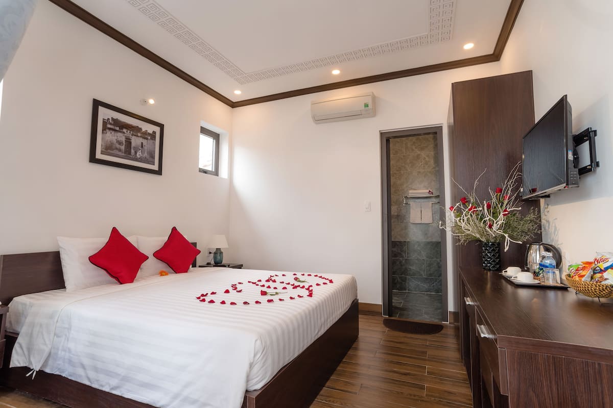 A hotel room with a neatly made bed, a television, a bathroom, and a wooden desk with a vase of flowers.