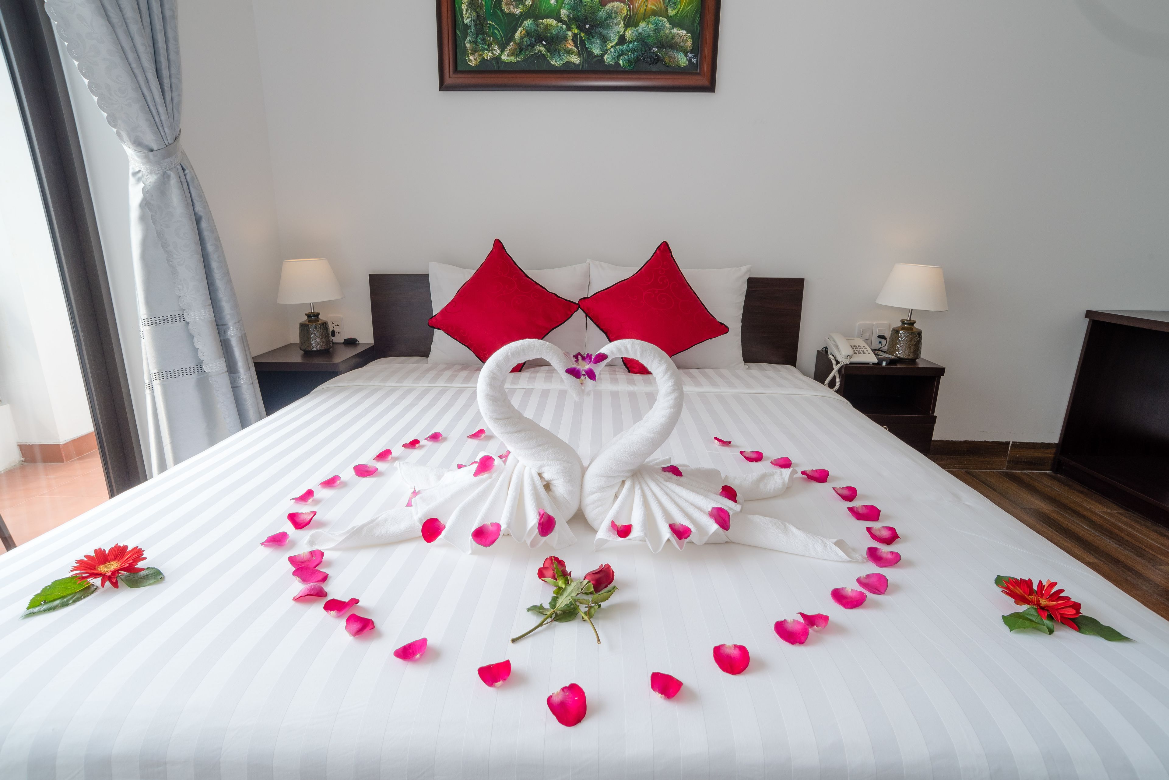 A neatly made bed with a heart-shaped arrangement of rose petals and swan-shaped towels, red pillows, and a single red flower on the bed.