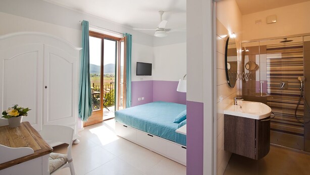 Un dormitorio con cama, televisión, balcón con cortinas, baño con lavabo y espejo, y una puerta.