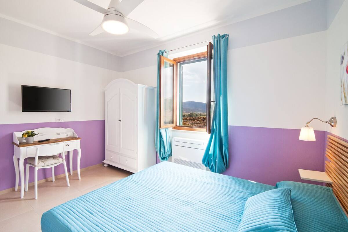 Een slaapkamer met een bed, een bureau met stoel, een televisie, een kledingkast en een raam met gordijnen.