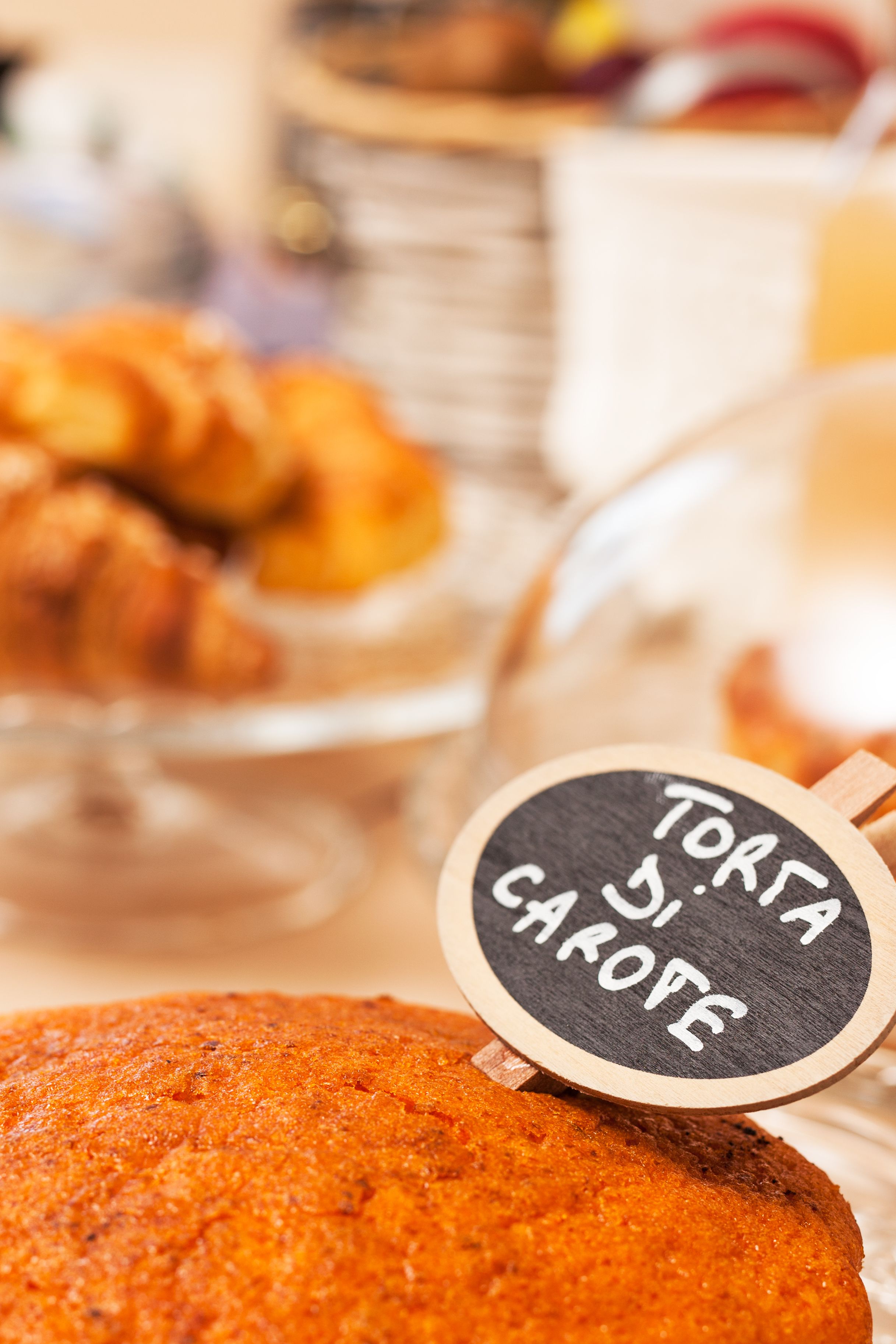 A round cake with a label reading "TORFA DI CAROTE" in front of a blurred background of food items.