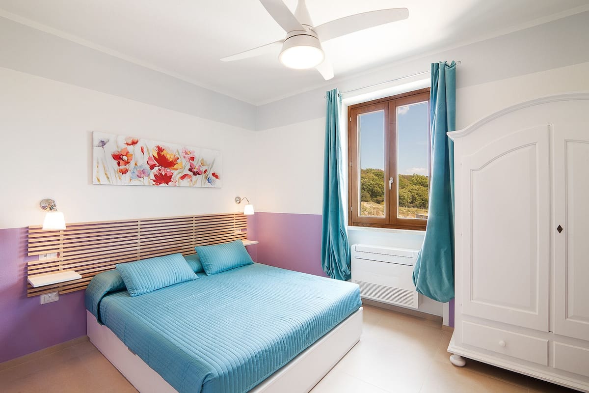Een slaapkamer met een bed, een plafondventilator, een raam met gordijnen, een kledingkast en een schilderij aan de muur.