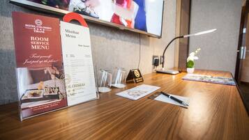 A hotel reception desk with a room service menu, a television displaying a scene, and a small table with a lamp and a vase of flowers.