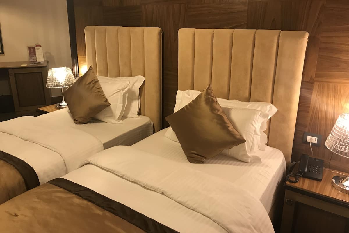 A hotel room with two beds, each with a headboard and pillows. There is a nightstand with a lamp and a phone.