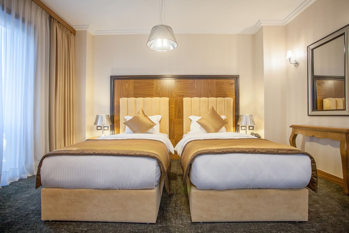 A hotel room with two beds, a wooden headboard, a hanging light fixture, a bedside table with a lamp, and a mirror on the wall.