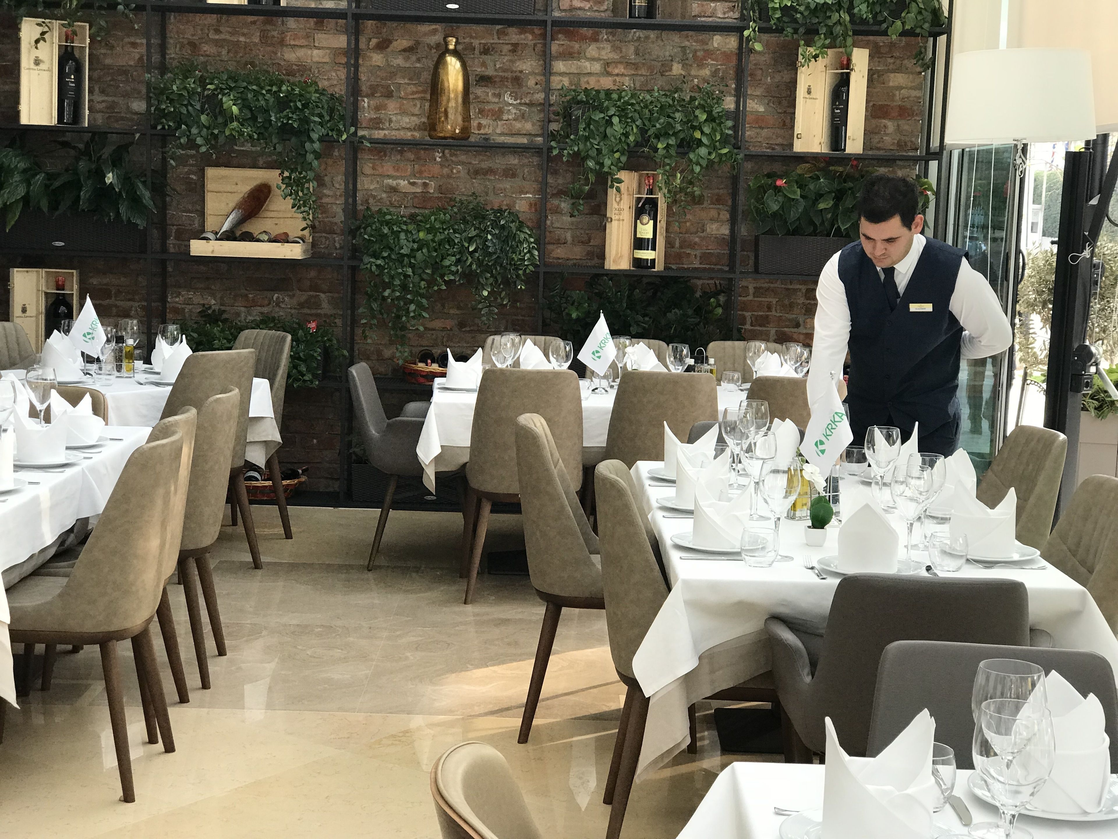 Un restaurante con mesas preparadas para comer, un camarero atendiendo una mesa, y una pared de ladrillo con plantas y botellas de vino.