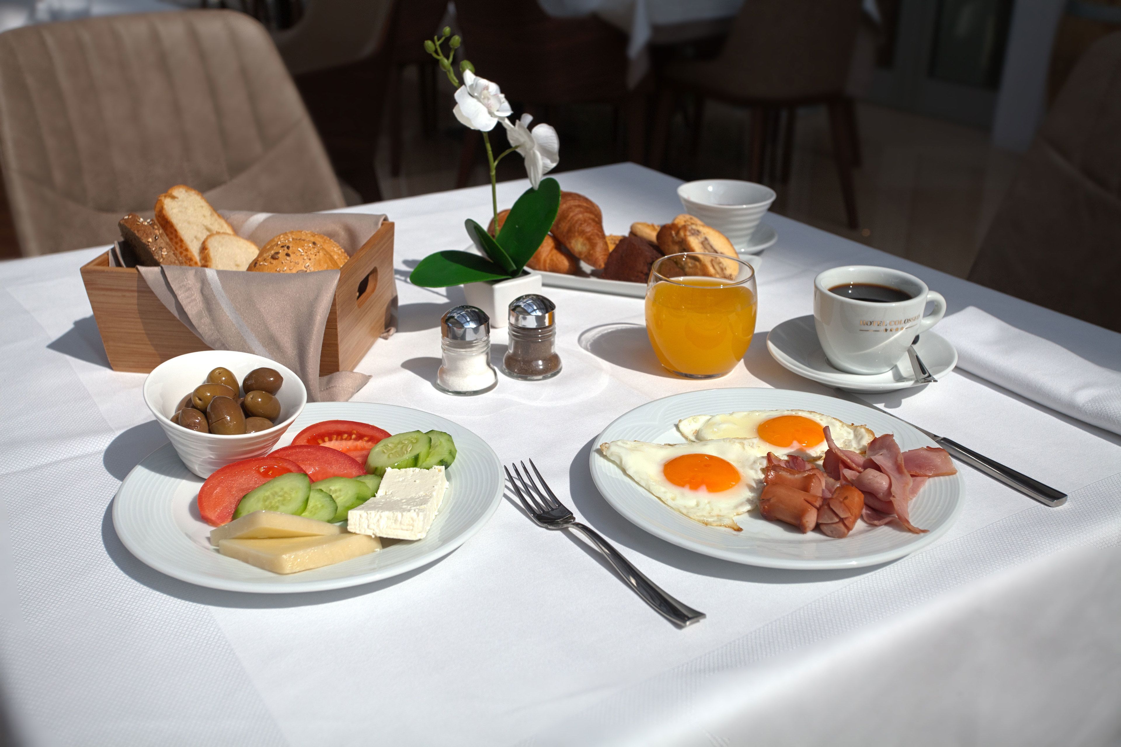 Una mesa de desayuno con cruasanes, huevos, jamón, queso, tomates, pepinos, aceitunas y una taza de café.