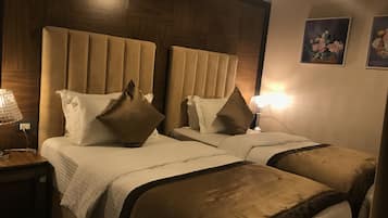 A hotel room with two beds, each with a headboard, white bedding, and brown throw pillows. There are bedside lamps and a wall-mounted picture.