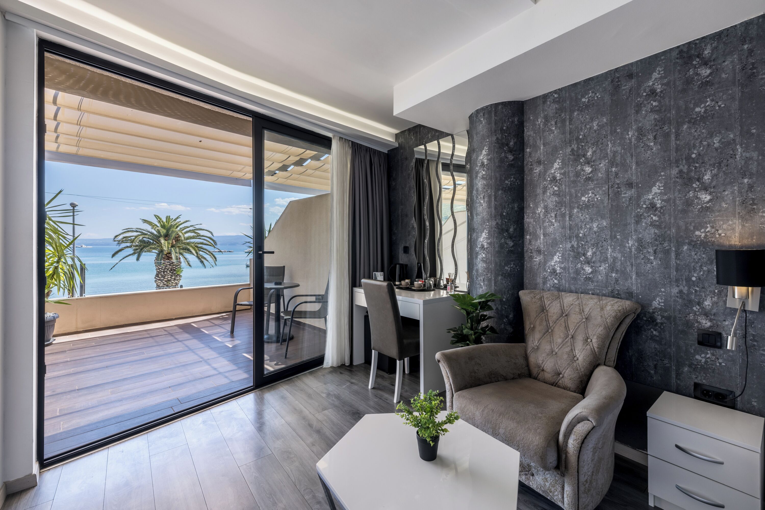 A modern living room with a large sliding glass door leading to a balcony, a tufted armchair, a white coffee table, and a desk with chairs.