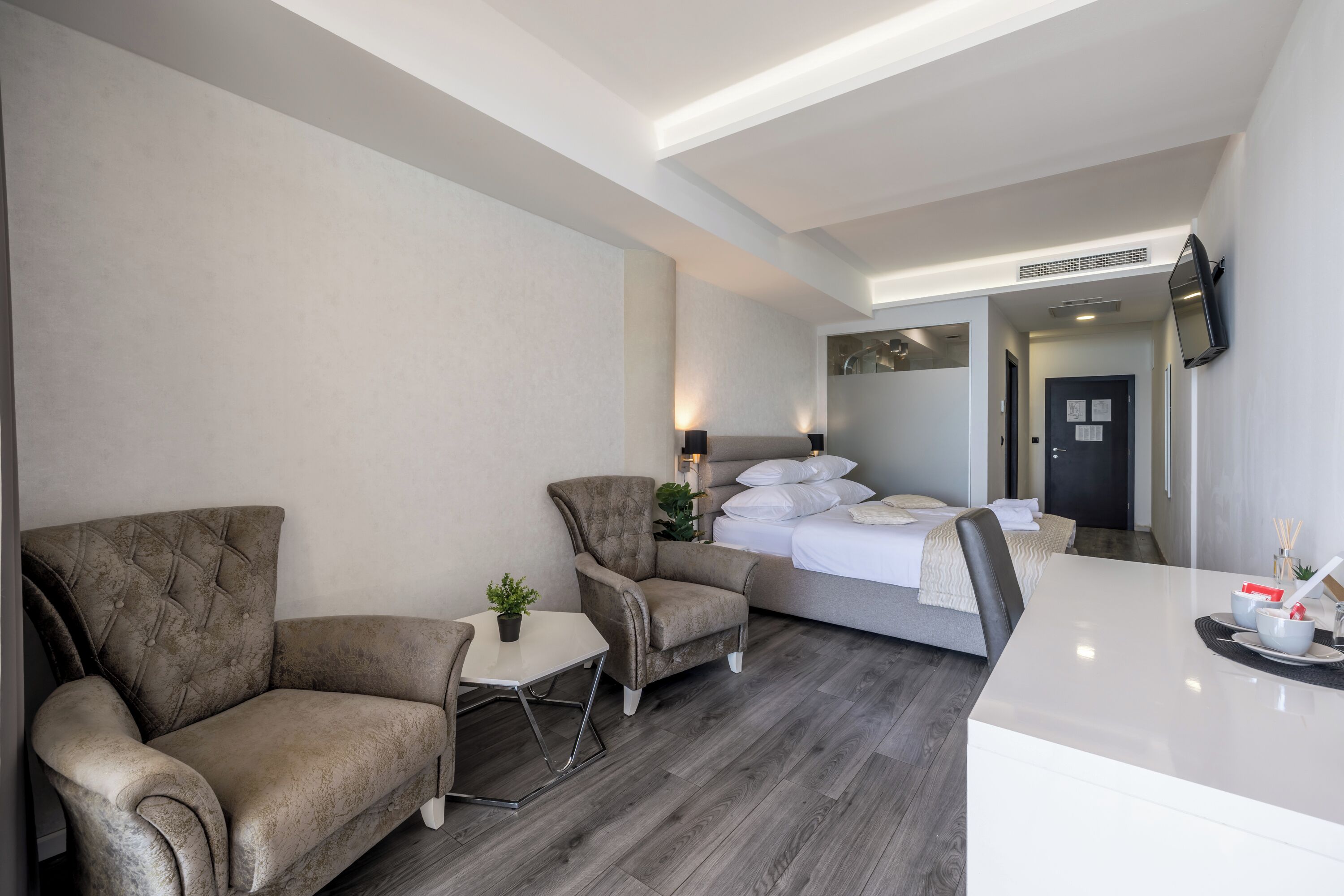 Une chambre d’hôtel moderne avec un canapé, une petite table agrémentée d’une plante, un lit avec du linge de maison blanc et un téléviseur à écran plat fixé au mur.