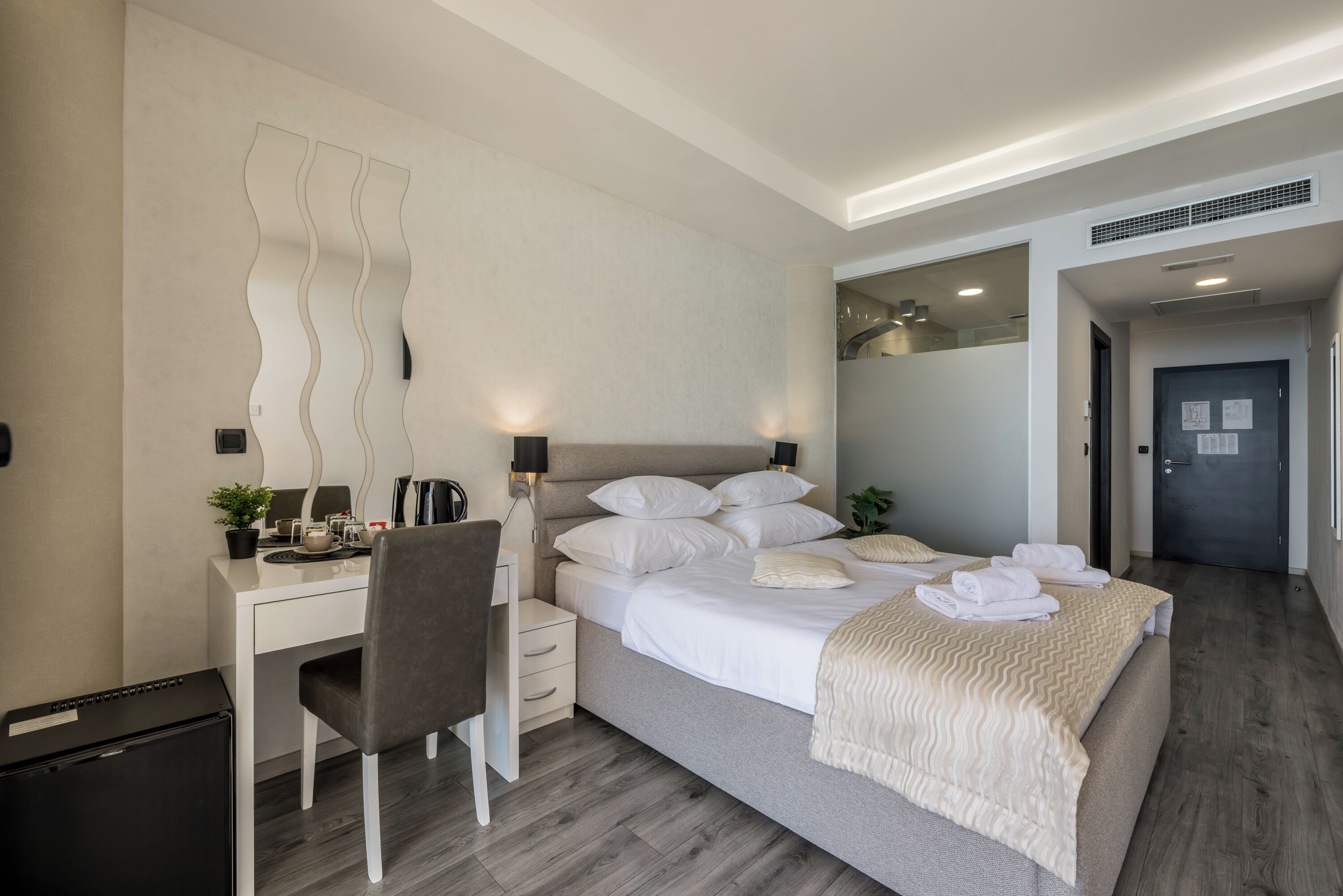 A modern hotel room with a bed, a desk, a chair, a small trash bin, and a wall-mounted mirror.