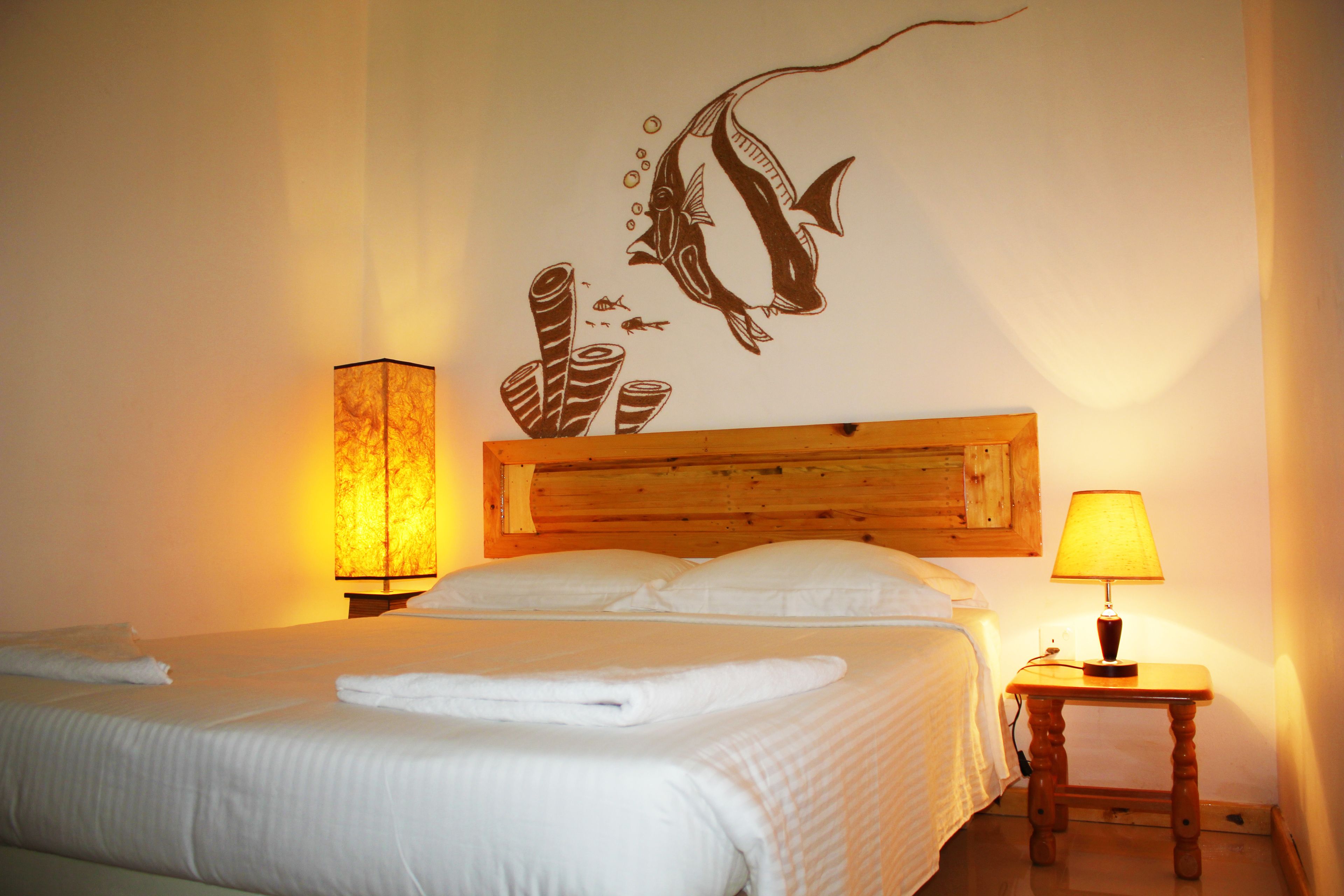 A neatly made bed with white linens and a wooden headboard, a bedside lamp, and a wall mural of a fish and coral.
