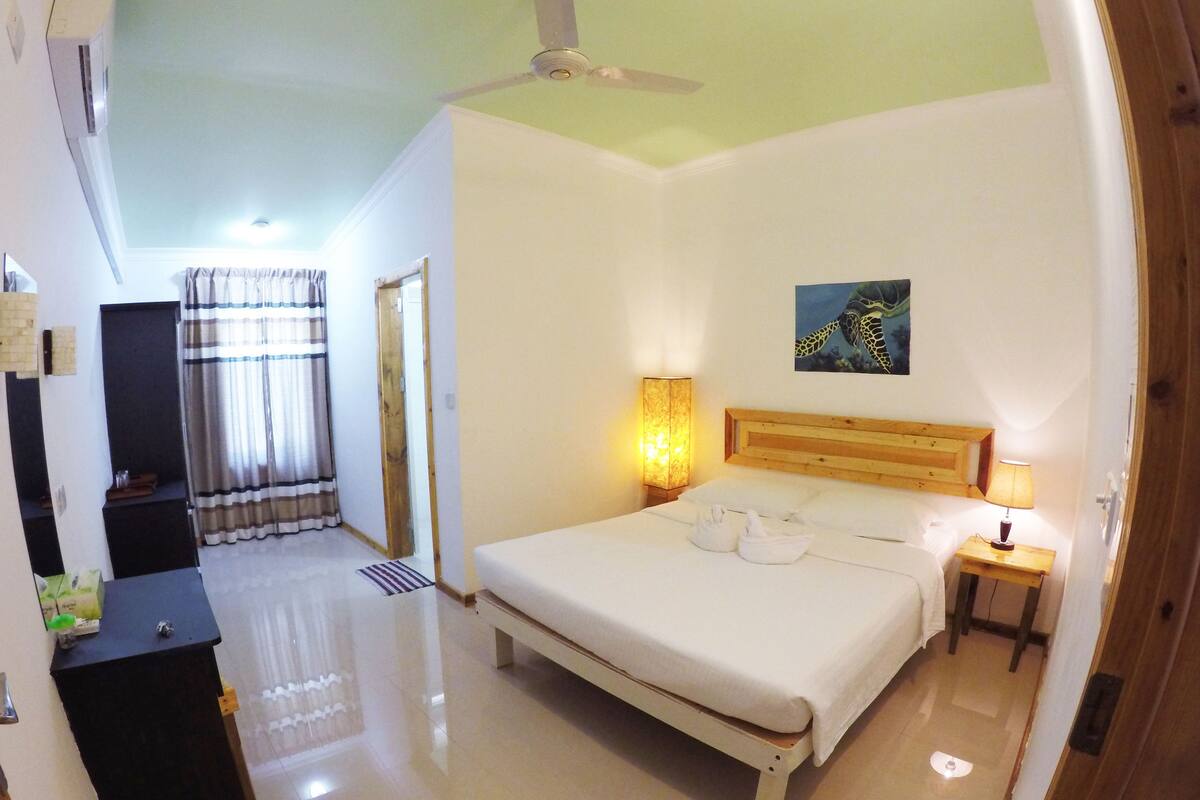 A hotel room with a bed, bedside lamps, a ceiling fan, a painting, and a bathroom visible through an open door.