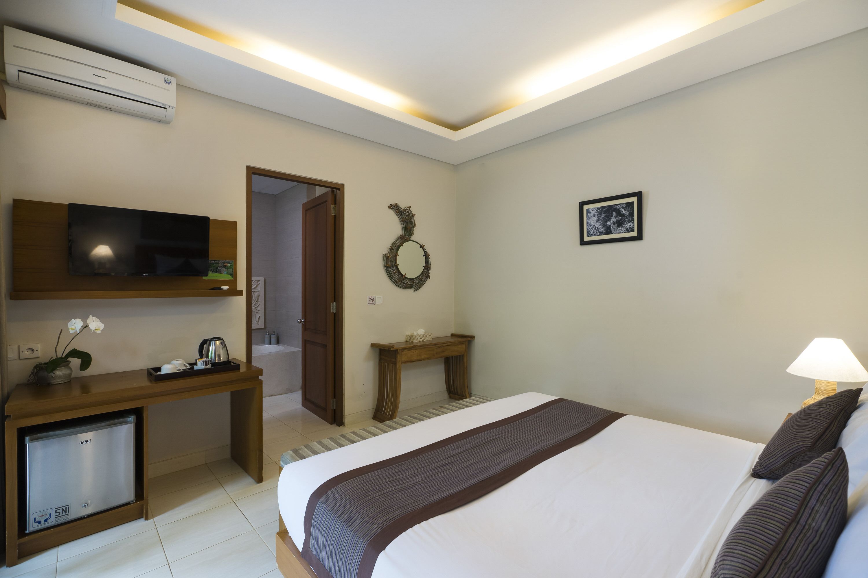 A hotel room with a bed, a desk with a television, a small refrigerator, a wooden console table, a wall-mounted air conditioner, a door, and a picture on the wall.