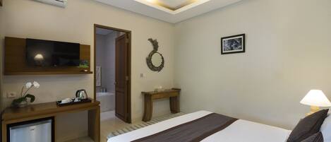 A hotel room with a bed, a desk with a television, a small refrigerator, a wooden console table, a wall-mounted air conditioner, a door, and a picture on the wall.