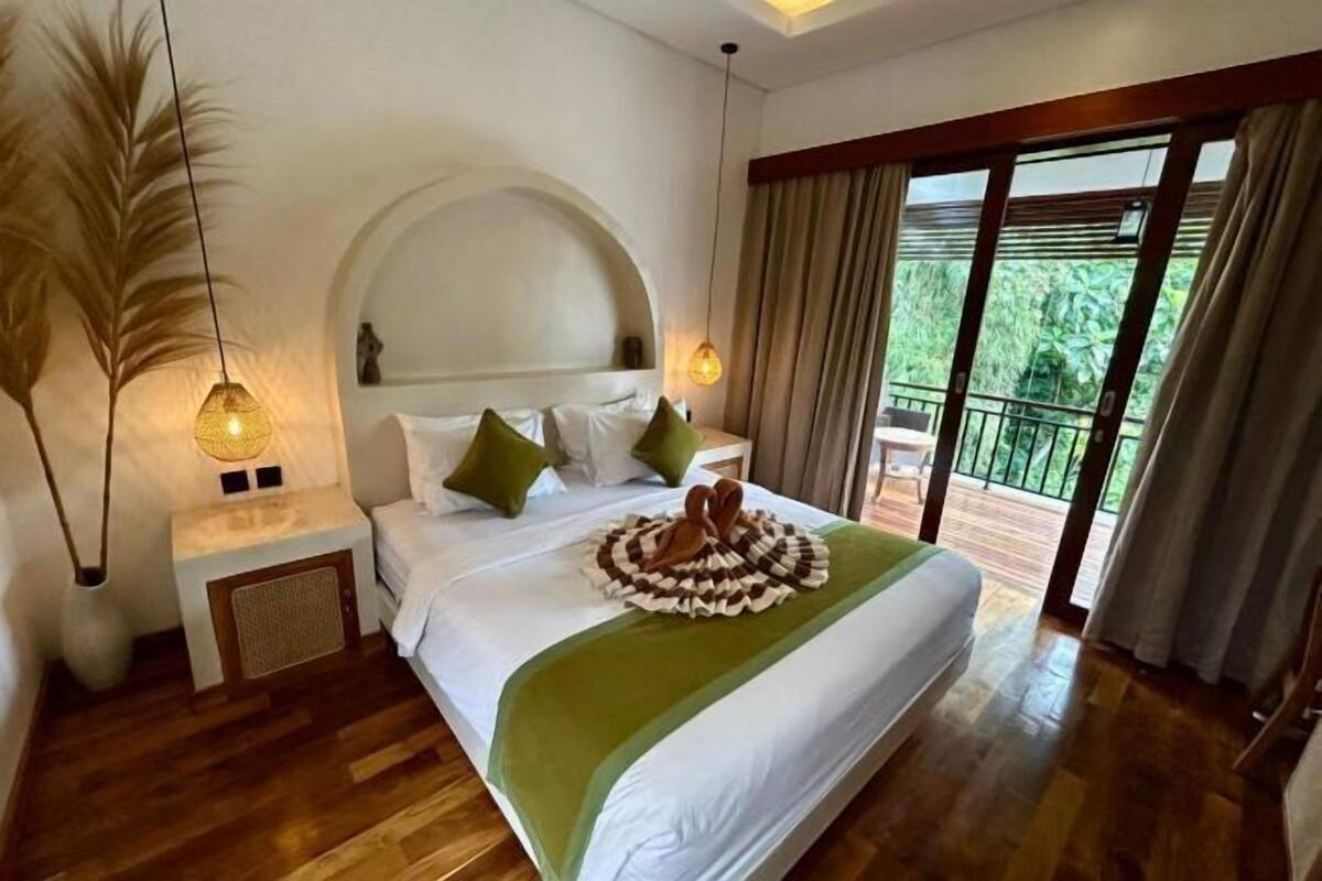 A hotel room with a bed, a nightstand, a lamp, a balcony with curtains, and a view of greenery.