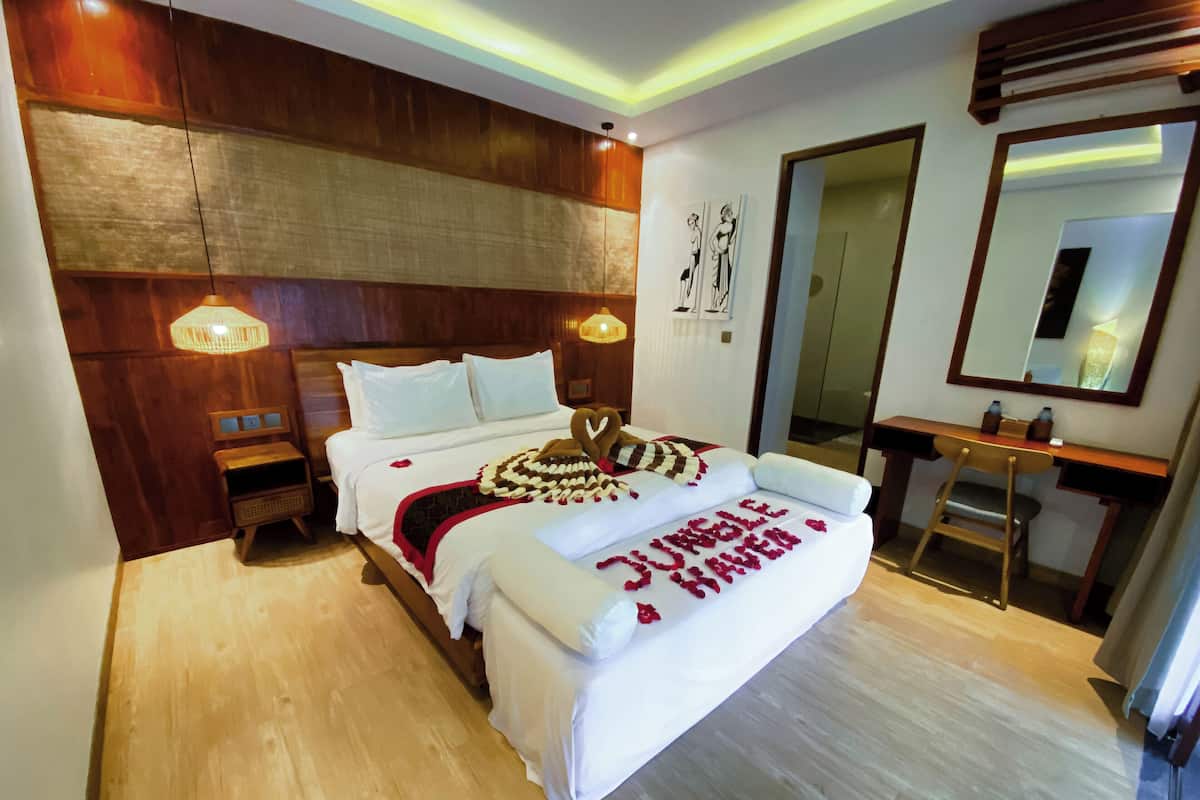 A hotel room with a wooden headboard, a bed with white linens, a bedside table with a lamp, a desk with a chair, and a large mirror on the wall.