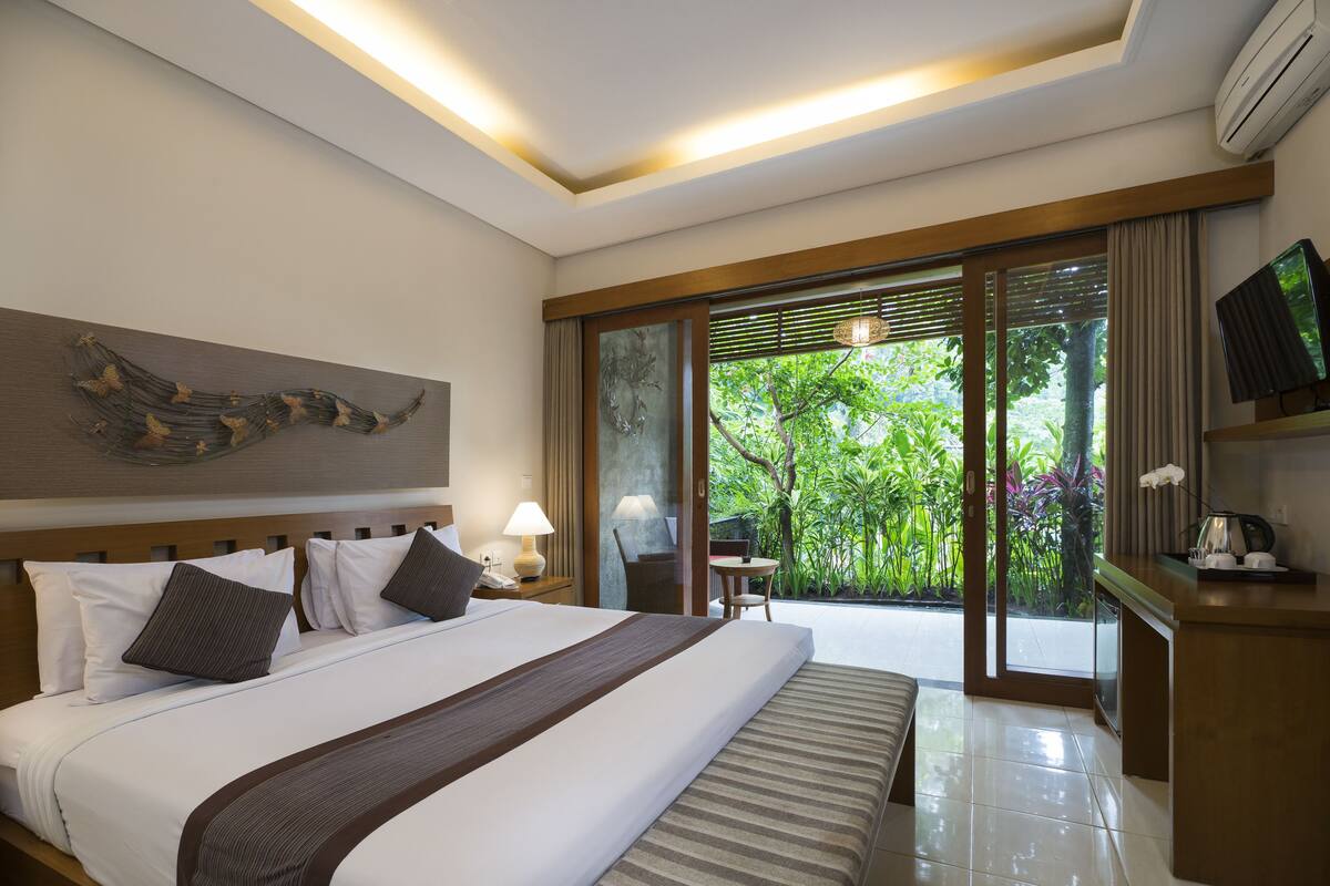 A hotel room with a large bed, a television, a desk with a lamp, and a view of greenery outside.