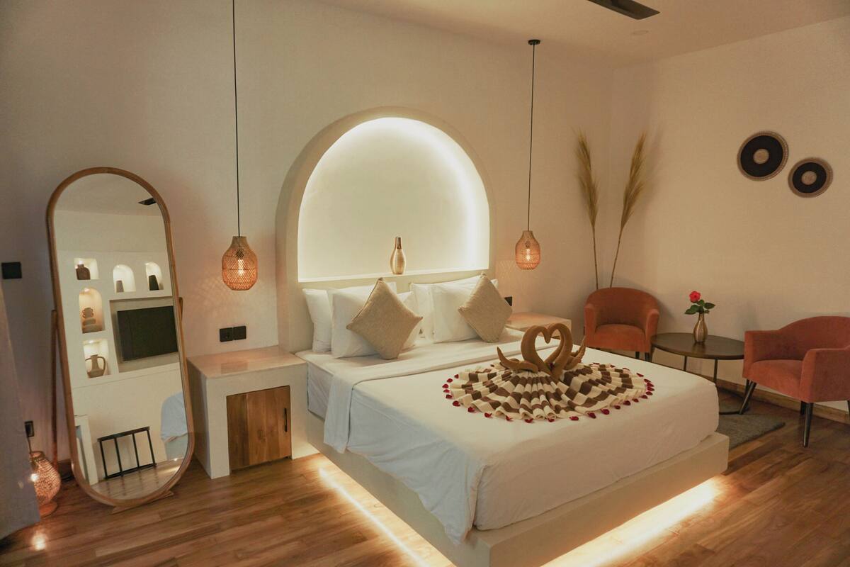 A modern bedroom with a large bed, a vanity with a mirror, two chairs, and a small table with a vase.