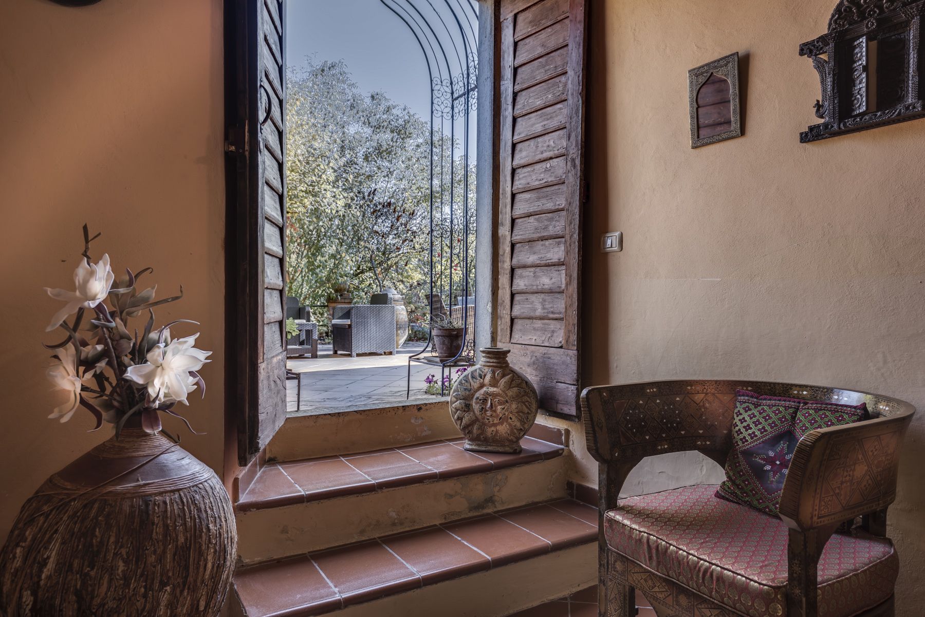 Une chaise en bois avec un coussin à motifs, un vase rempli de fleurs blanches et une vue sur un patio extérieur agrémenté de plantes en pot et d’un portail en fer forgé.