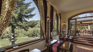 Un comedor con mesas y sillas, ventanas amplias con cortinas y vista a árboles y un césped.