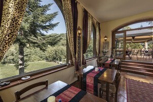 Un comedor con mesas y sillas, ventanas amplias con cortinas y vista a árboles y un césped.
