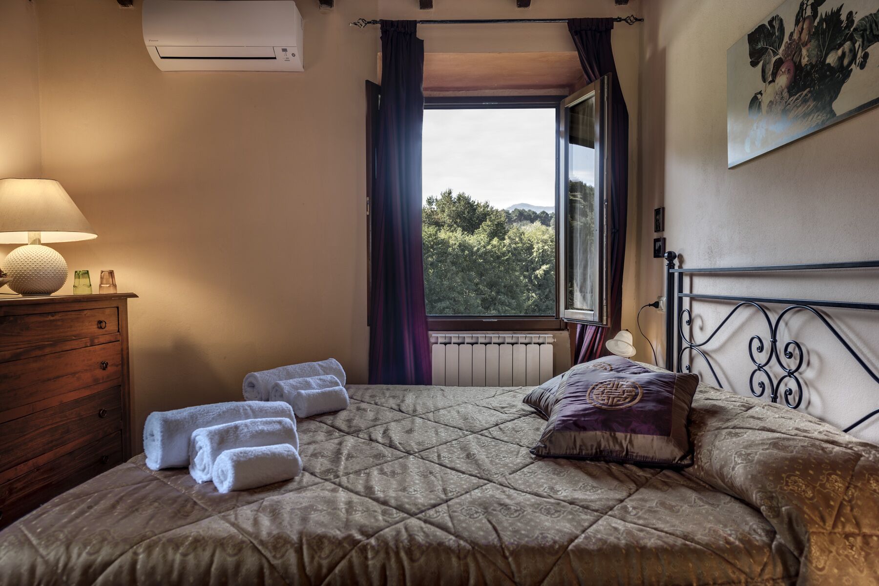 A bedroom with a bed, a nightstand, a lamp, a window with a view of trees, and an air conditioning unit.