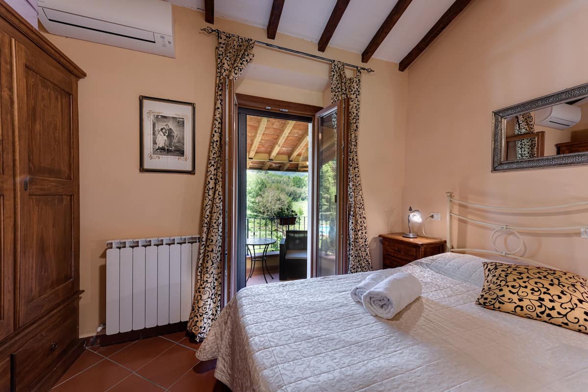 A bedroom with a bed, a bedside table with a lamp, a radiator, a wooden wardrobe, a window with curtains, and a view of an outdoor patio.