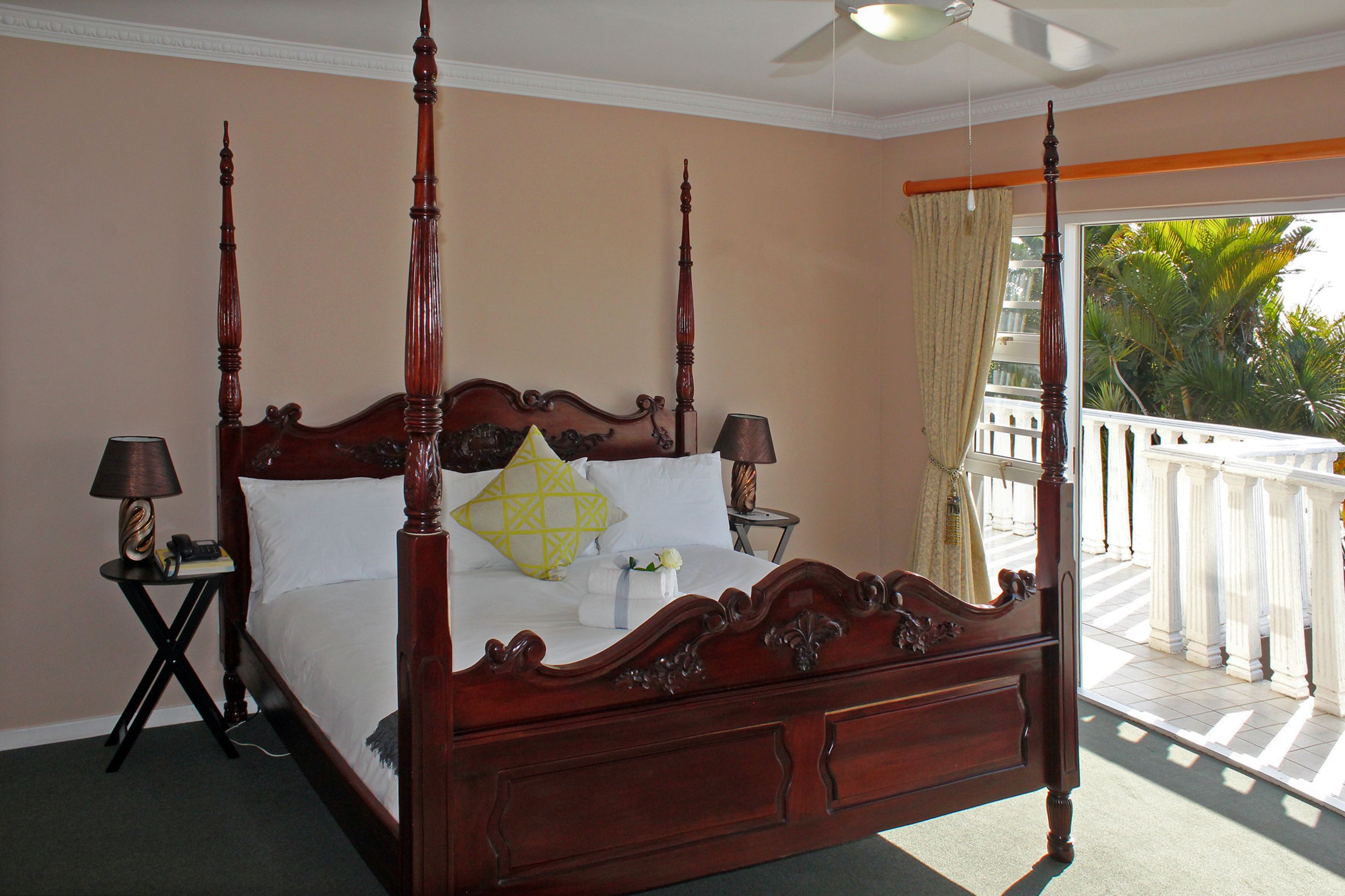 A four-poster bed with a wooden headboard, two bedside tables with lamps, and a view of a balcony with a white railing and palm trees.