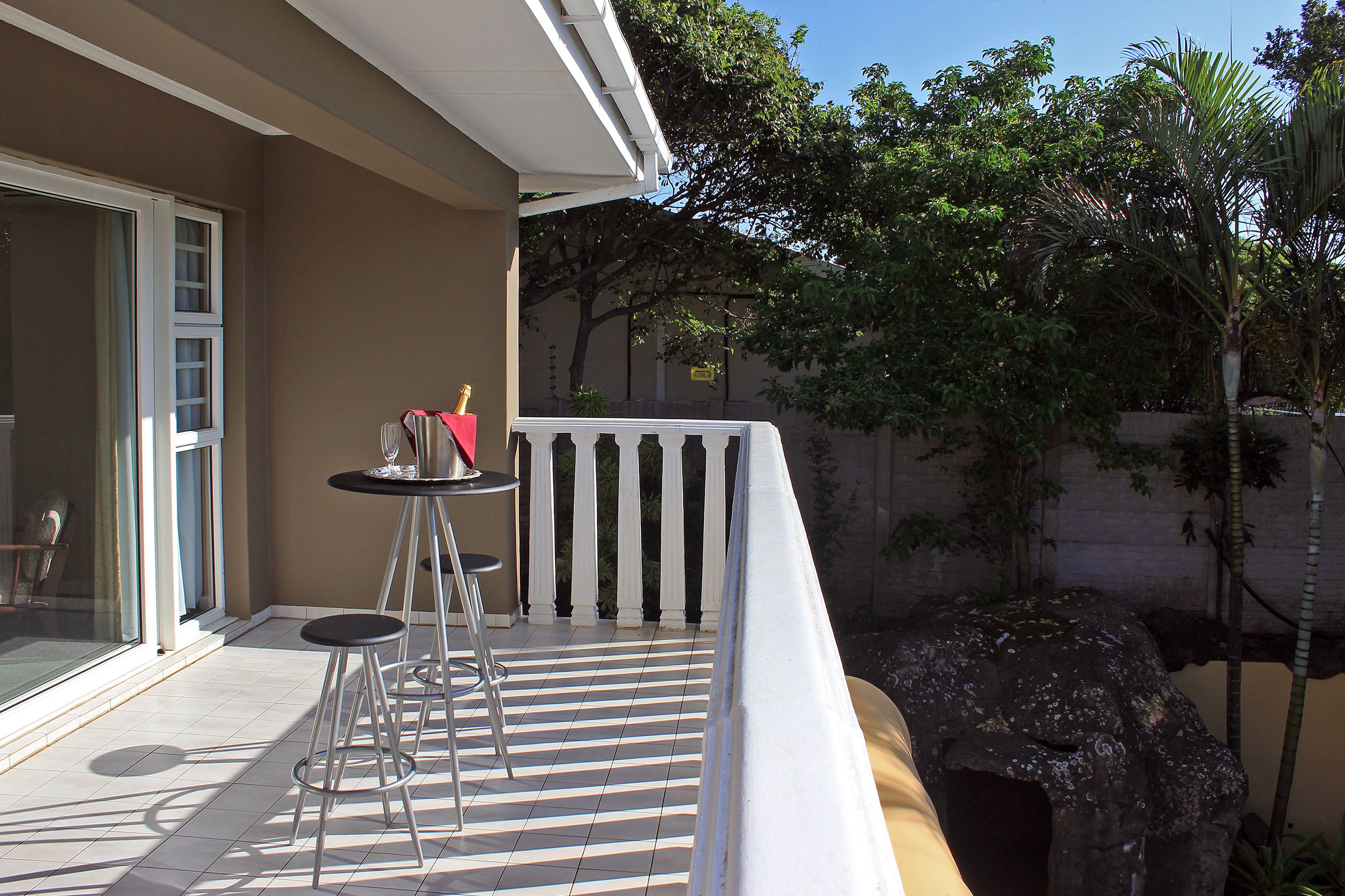 A balcony with a tiled floor, two bar stools, a small table with a bottle, and a decorative rock.