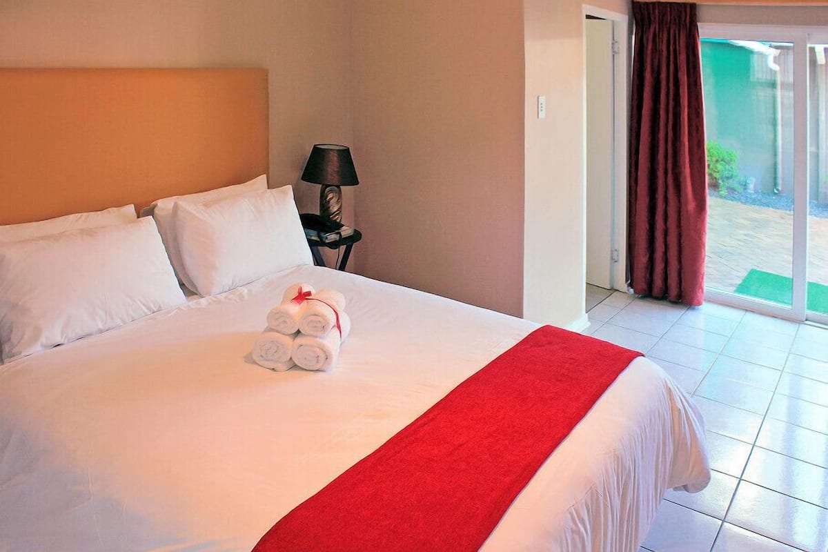 Uma cama bem arrumada com lençóis brancos e um cobertor vermelho, um abajur em uma mesa de cabeceira, e vista para uma área verde externa através de uma porta deslizante.