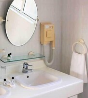 Un baño con lavamanos blanco, estante de vidrio con artículos de aseo, espejo y argolla para toallas.