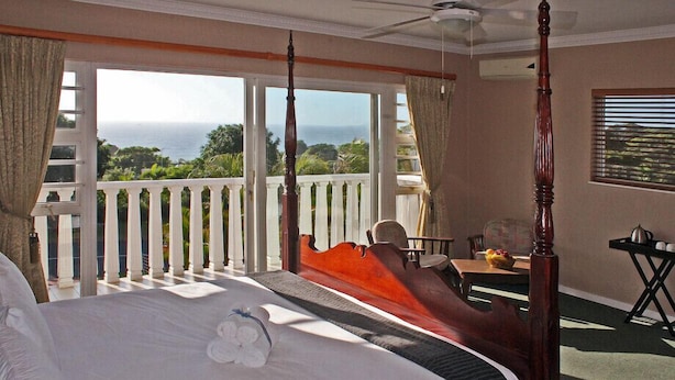 Un lit à baldaquin avec vue sur l’océan, un balcon avec une balustrade blanche et un coin repas avec une table et des chaises.