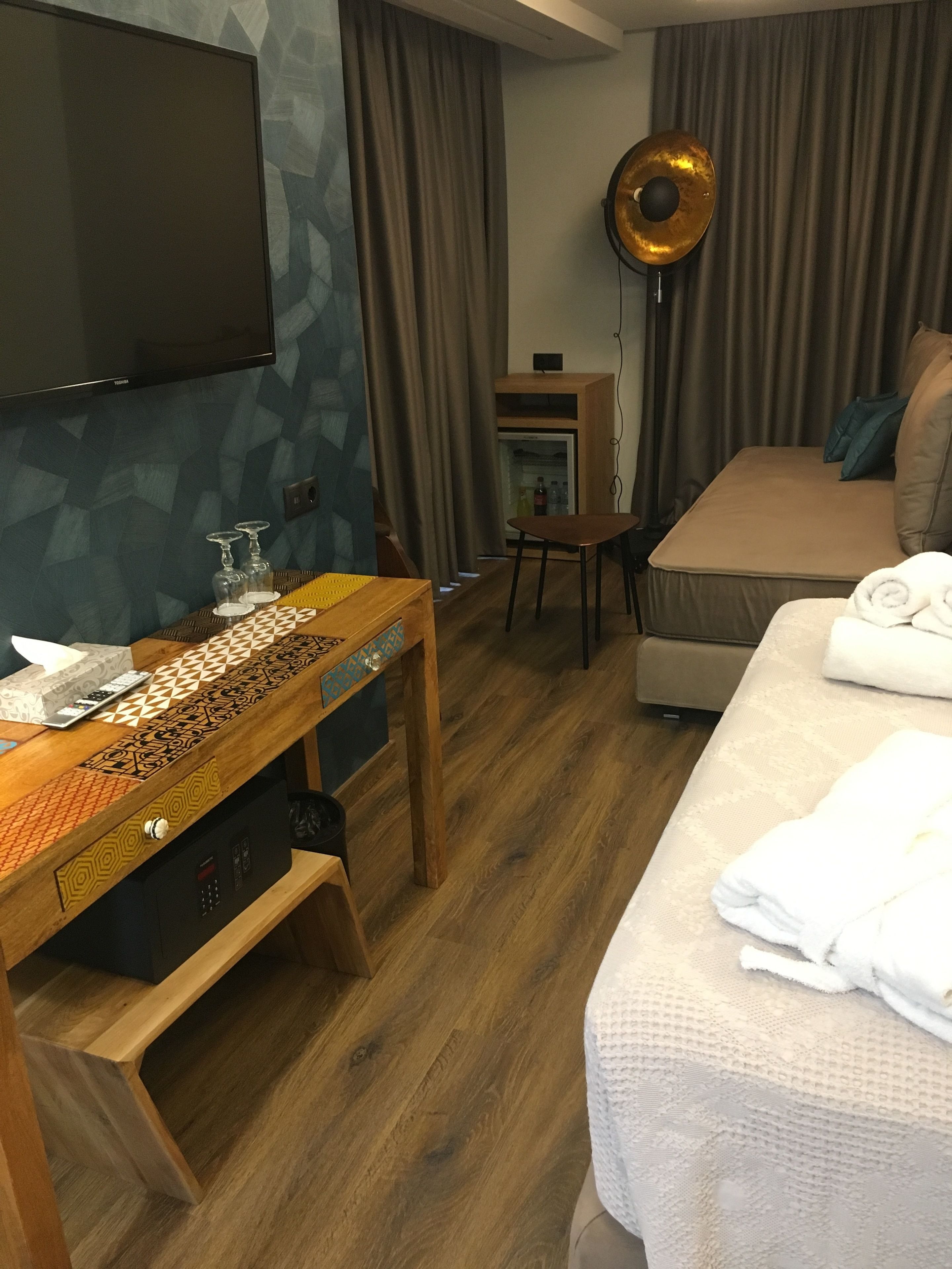 A hotel room with a bed, a wooden table with a patterned design, a television mounted on the wall, a small cabinet, and a round floor lamp.