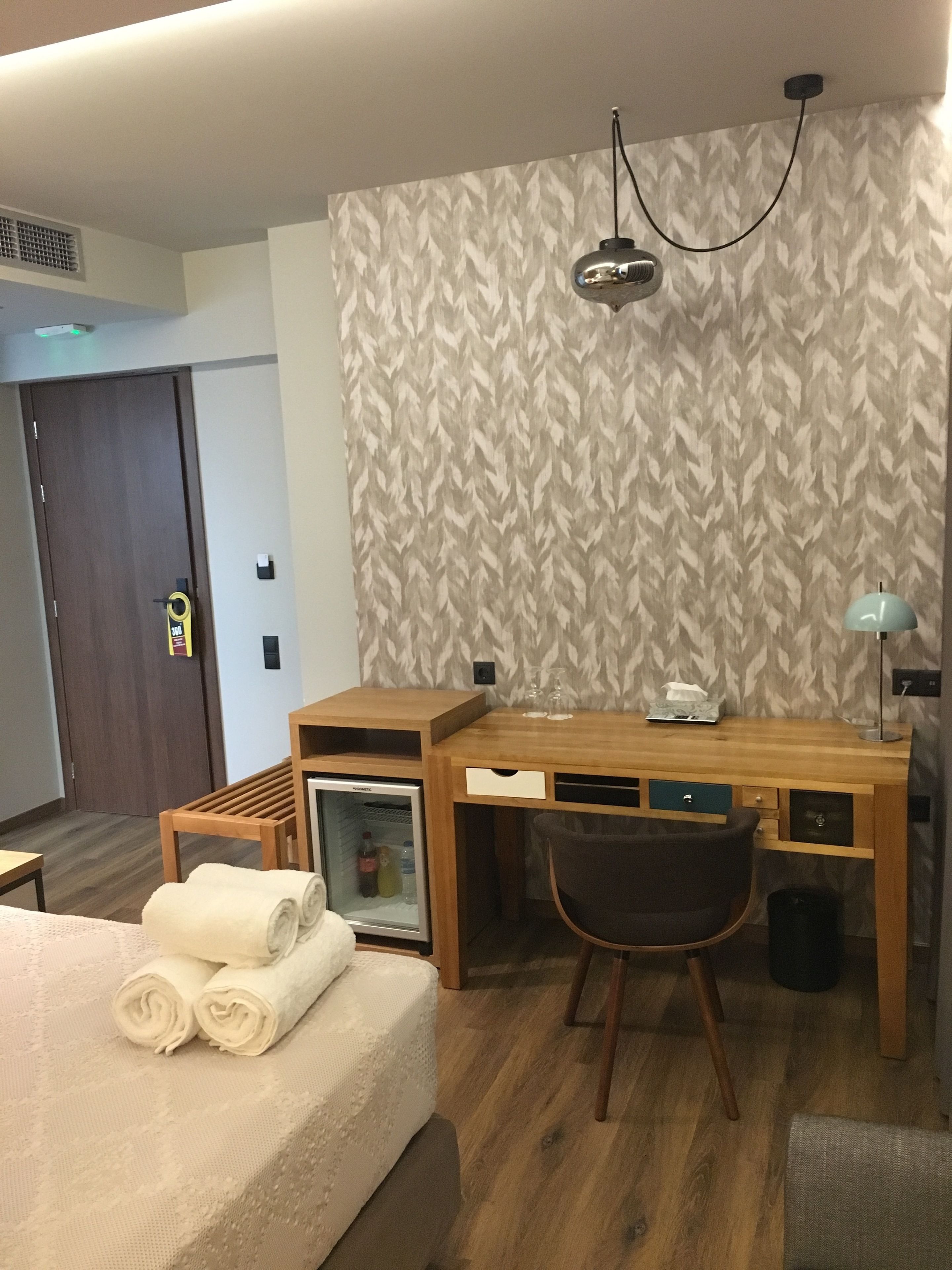 Hotellihuone, jossa on puinen työpöytä, tuoli, sängyllä rullalle taiteltuja pyyhkeitä, minibaari ja kuviollinen tapetti.