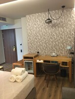 Hotellihuone, jossa on puinen työpöytä, tuoli, sängyllä rullalle taiteltuja pyyhkeitä, minibaari ja kuviollinen tapetti.