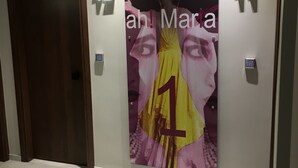 A hallway with a marble floor, a wooden door, and a wall with a large poster featuring a person in a yellow dress.