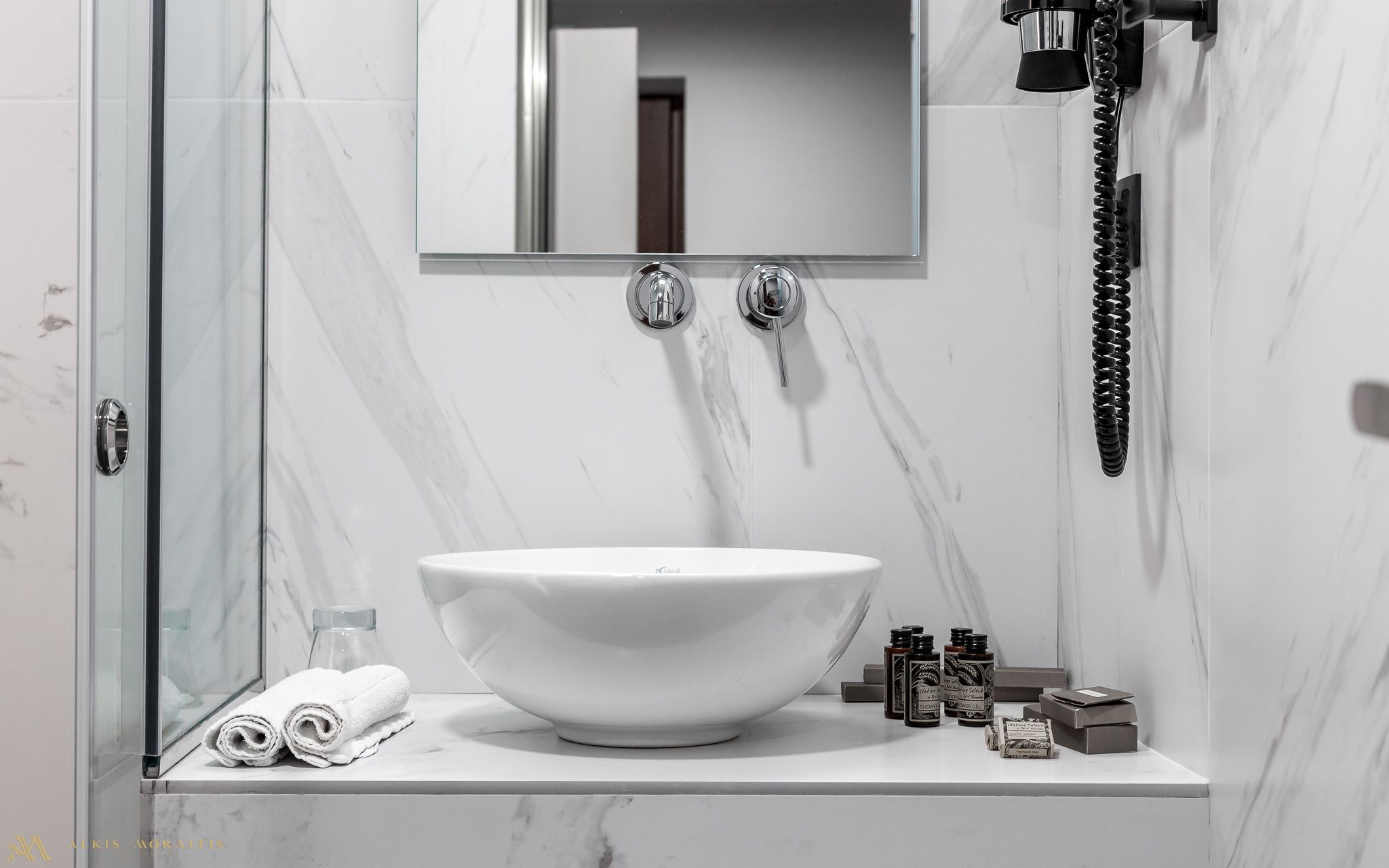 A bathroom with a large white vessel sink, a glass shower enclosure, and a marble-tiled wall.