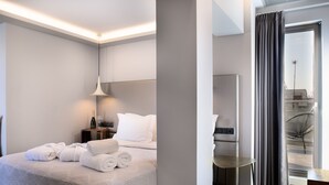 A modern hotel room with a neatly made bed, a bedside table with a bottle and glasses, and a view of the outside through a large window with curtains.