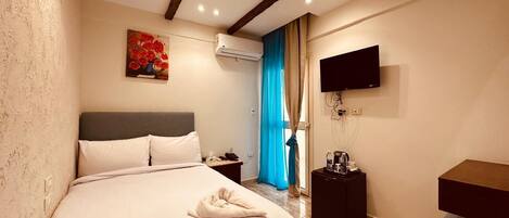 A hotel room with a bed, bedside table, air conditioning unit, TV, and a painting on the wall.