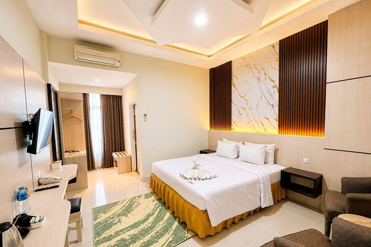 A hotel room with a bed, bedside tables, a desk, a chair, a TV, and an air conditioning unit.