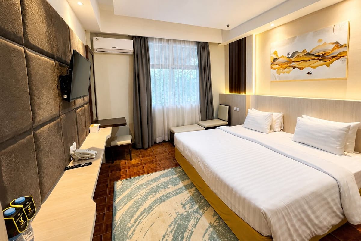 A hotel room with a large bed, a desk with a chair, a flat-screen TV, an air conditioning unit, and a painting on the wall.
