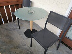 Een ronde glazen tafel met een metalen onderstel, een enkele stoel met een net rugleuning en een houten hek op de achtergrond.