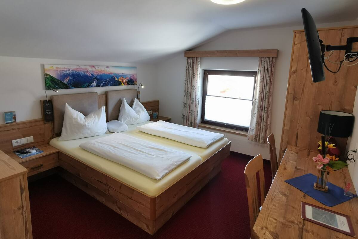 A bedroom with a wooden bed, a desk, a chair, a window with curtains, a painting on the wall, and a ceiling light.